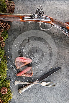 Deer or venison steak with antique long gun, cutlery and ingredients like sea salt and pepper, food background for restaurant or h