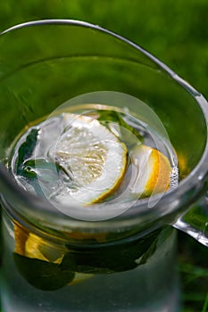 Defocus close-up lemon and mint in glass jug of lemonade on natural green nature background. Pitcher of cold summer