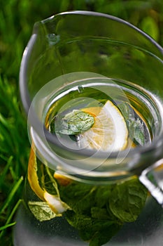 Defocus close-up slice lemon and leaves of mint in glass jug of lemonade natural green background. Pitcher of cool