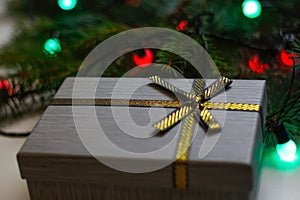 Defocus gray gift with close-up gold bow ribbon on pine or fir tree blurred background with glowing festive light bulb