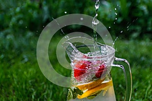 Defocus jet splash spray water bubbles in glass jug of lemonade with lemon, strawberry and mint on natural green