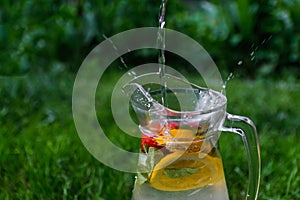 Defocus jet splash spray water bubbles in glass jug of lemonade with lemon, strawberry and mint on natural green