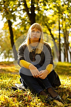 Defocus serious blond 40s woman seating in yellow autumn park. Happy beautiful lady. Women wearing grey pullover, yellow