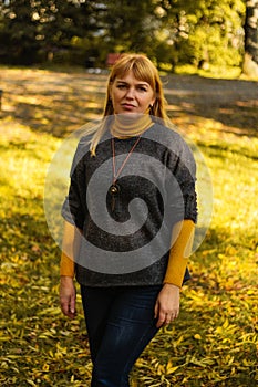 Defocus serious blond 40s woman standing in yellow autumn park. Happy beautiful lady. Women wearing grey pullover