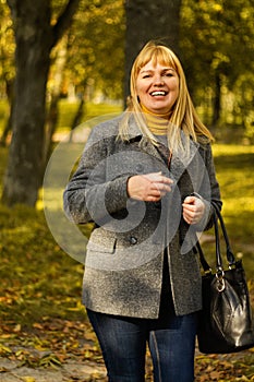 Defocus smiling blond 40s woman gesturing in yellow autumn park. Happy beautiful lady. Women wearing grey coat, pullover