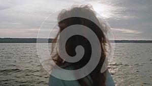 Defocused back view of brunette with long hair standing on pier in front of water. Sky and water on background are in