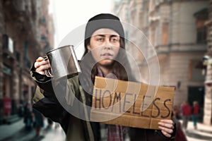 Defocused beggar woman holds a cardboard sign with the text homeless and steel alms cup. Defocused background with