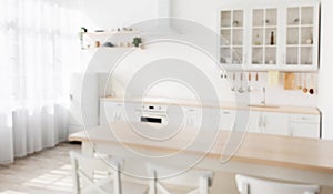 Defocused photo of wooden dining table and white chairs nearby, various utensils on light kitchen furniture