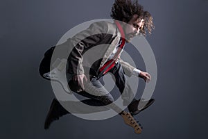 Defocused studio portrait of a young bearded, curly-haired guy with long hair, in a jacket, jumping with a guitar, looking at the