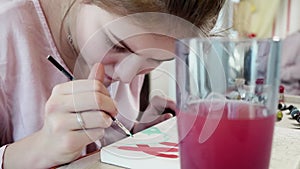 Defocused view of glass of water on foreground and focused view of blond girl colouring details of picture in mint