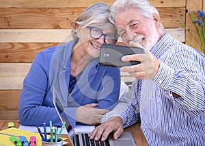 Defocused white-haired elderly people making a selfie with a black smart phone while share the same wooden table to work from home