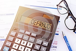 Defraud is a word written on calculator, lying on white surface next to glasses and a blue pen photo
