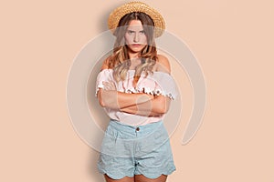 Dejected European woman with dissatisfied expression, keeps hands crossed, wears summer straw hat, fashionable blouse and shorts,