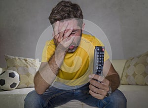 Dejected and frustrated man watching European football game on TV sitting at living room couch alone feeling sad and unhappy with