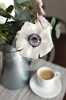 Delicate anemones flowers in metal pot vase with cup of coffee on the table