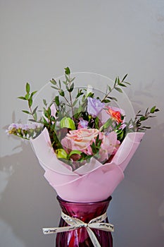 A delicate bouquet of different flowers in a light pink wrapper stands in a burgundy vase on a gray background