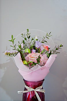 A delicate bouquet of different flowers in a light pink wrapper stands in a burgundy vase on a gray background