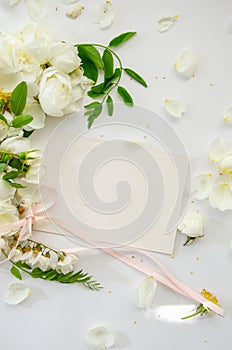 Delicate composition of blank card with white roses and petals with acacia flowers and pink ribbon on white background. Wedding