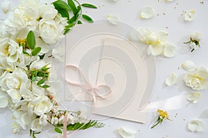 Delicate composition of blank card with white roses and petals with acacia flowers and pink ribbon on white background. Wedding