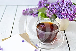 Delicate morning tea table setting with lilac flowers, a transparent cup of tea and a saucer and a white vase, white and