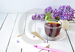 Delicate morning tea table setting with lilac flowers, a transparent cup of tea and a saucer and a white vase, white and