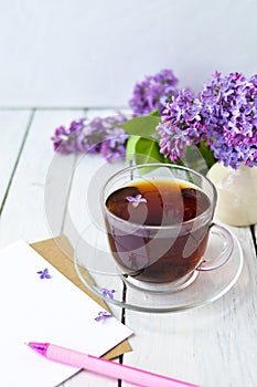 Delicate morning tea table setting with lilac flowers, a transparent cup of tea and a saucer and a white vase, white and