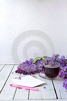 Delicate morning tea table setting with lilac flowers, a transparent cup of tea and a saucer and a white vase, white and