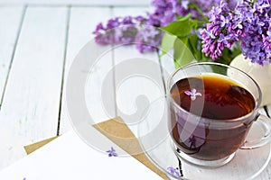 Delicate morning tea table setting with lilac flowers, a transparent cup of tea and a saucer and a white vase, white and craft