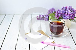 Delicate morning tea table setting with lilac flowers, a transparent cup of tea and a saucer and a white vase, white and craft