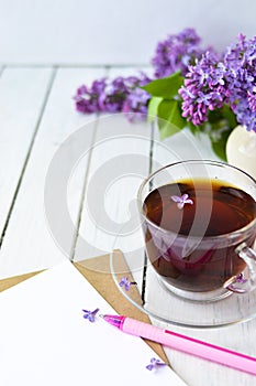 Delicate morning tea table setting with lilac flowers, a transparent cup of tea and a saucer and a white vase, white and craft