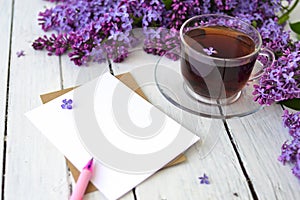 Delicate morning tea table setting with lilac flowers, a transparent cup of tea and a saucer and a white vase, white and craft