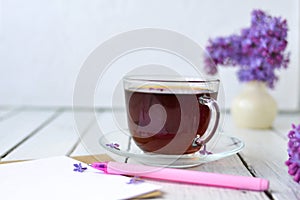 Delicate morning tea table setting with lilac flowers, a transparent cup of tea and a saucer and a white vase, white and craft