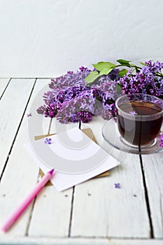 Delicate morning tea table setting with lilac flowers, a transparent cup of tea and a saucer and a white vase, white and craft