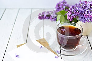 Delicate morning tea table setting with lilac flowers, a transparent cup of tea and a saucer and a white vase, white and craft