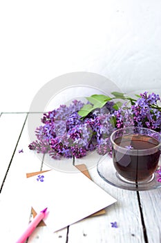 Delicate morning tea table setting with lilac flowers, a transparent cup of tea and a saucer and a white vase, white and craft