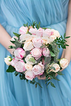Delicate wedding bouquet of pink and cream roses in the bride`s hands on a background of light blue chiffon dress close-up.