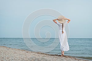 Delicate young woman in white dress and hat walks on sea shore. Back view