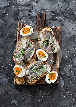 Delicious appetizer, tapas - soft boiled eggs and canned sardines sandwiches on a cutting board on a dark background, top view