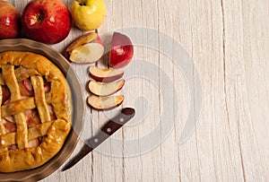 Delicious Apple pie in ceramic form, knife, sliced red apples on a light wooden background.