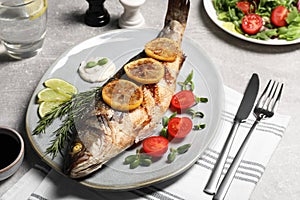 Delicious baked sea bass fish served with lemon, tomatoes and sauce on light grey table
