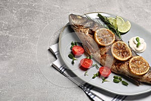 Delicious baked sea bass fish served with lemon, tomatoes and sauce on light grey table. Space for text