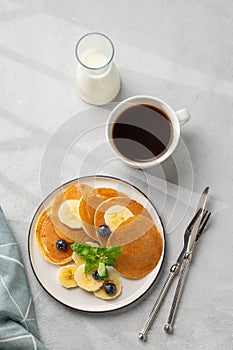 Delicious banana pancakes with fresh fruit slices, berries, mint and honey on a light background with a cup of black coffee, soy