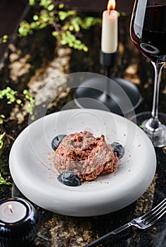 Delicious beef meat tartare with pickled cucumber and truffle sauce on plate on marble table with wine glasses. Healthy food,