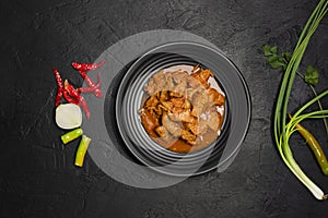 Delicious beef or pork stew in a rustic black plate with fresh coriander, red chilies, garlic, onions and spices placed on a black