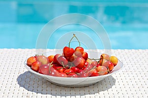 A delicious bowl of bright red and yellow cherries sitting on the table poolside.
