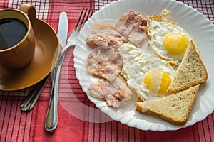 Delicious Breakfast - a Cup of coffee, a plate of fried eggs, bacon and toast, next to the Cutlery on red checkered napkin