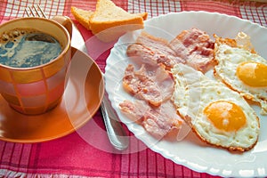 Delicious Breakfast - a Cup of coffee, a plate of fried eggs, bacon and toast, next to the Cutlery on red checkered napkin