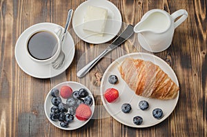Delicious breakfast with fresh berries. Coffee cup, creamer and croissant with berries in white bowl and butter on wooden table.