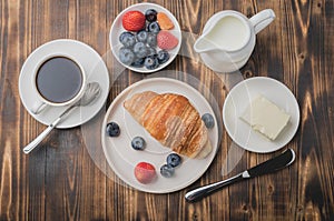 Delicious breakfast with fresh berries. Coffee cup, creamer and croissant with berries in white bowl and butter on wooden table.