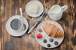 Delicious breakfast with fresh berries. Coffee cup, creamer and croissant with berries in white bowl and butter on wooden table.